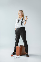 attractive fashionable girl smoking cigar while standing on grey with vintage travel bag, newspaper...