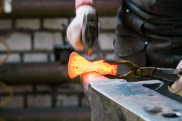 Blacksmith working with red hot metal workpiece of new axe on the anvil at the forge. Focus on red hot metal
