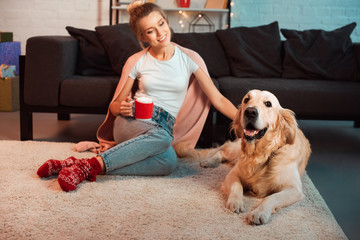 Happy young blonde woman dog sitting on fluffy rug with cup of hot cocoa and stroking golden retriever dog at Christmas time