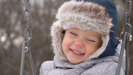 Smiling kid swaying on swing. Cute little boy (toddler, two year old) in hat with fur trim and earflaps.  Happy child. Winter, cold weather, outdoor