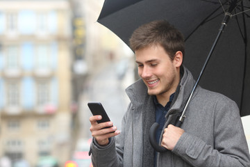 Happy man checking phone under an umbrella in winter