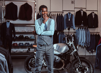 Obraz na płótnie Canvas Elegantly dressed African American young man posing near retro sports motorbike at the men's clothing store.