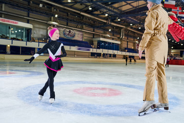 Full length portrait of happy little girl figure skating in indoor rink and looking at female coach, copy space