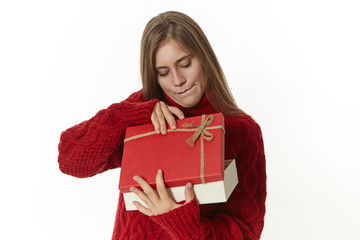 Curious female in knitted warm sweater holding fancy red box, opening it impatiently, looking inside with interest. Pretty girl receiving birthday gift from boyfriend. Celebration, gifts and presents