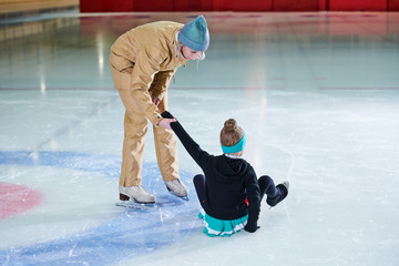 Full length portrait of female coach helping little girl get up during figure skating training in...