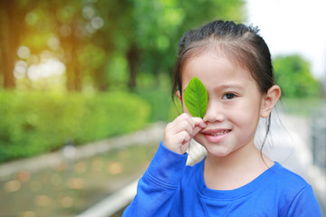 Fototapeta premium Adorable little Asian child girl holding a green leaf closing right eye in green garden background.