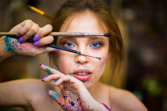 Close up portrait of beautiful female artist with dirty hands with different paints on them, holding paint brushes near her face and eyes.