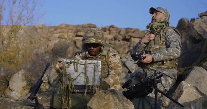 Medium shot of fully equipped and armed african american soldier looking at a computer and another talking on a radio and launches exploration drone