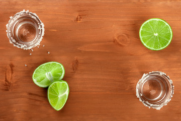 A photo of two tequila shots with limes, shot from above on a dark rustic wooden background, forming a frame for copy space