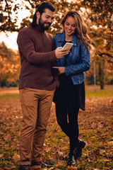 Fototapeta premium Young couple using cellphone in autumn colored park.
