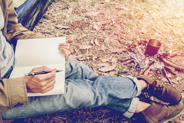 Asian young man sitting is reading and writing a book in outside the tent. Alone camping in forest.
