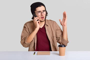 Emotional displeased guy has telephone coversation, raises hand, discusses something actively, drinks takeaway coffee, sits at desktop against white background. People and technology concept