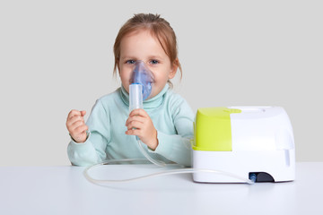Pretty small child uses nebulizer mask for inahlation, cares of health, sits at white desktop,...
