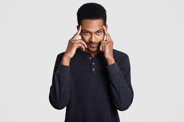 Contemplative dark skinned man keeps both index fingers on temples, thinks about something, tries to concentrate and remember some information, dressed in casual clothes, isolated over white wall