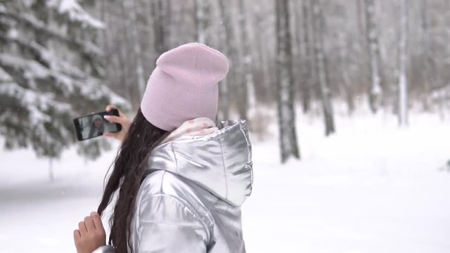 Attractive girl makes selfie using a smartphone while standing in a winter forest. 4K
