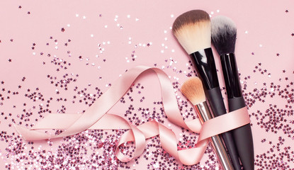 Different Cosmetic makeup brushes with pink ribbon and holographic glitter confetti in the form of...