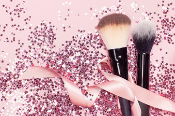 Two different Cosmetic makeup brushes with pink ribbon and holographic glitter confetti in the form of stars on pink background Flat lay top view copy space Makeup accessories holiday birthday newyear