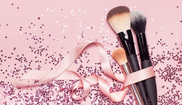Different Cosmetic makeup brushes with pink ribbon and holographic glitter confetti in the form of stars on pink background Flat lay top view copy space. Makeup accessories holiday birthday new year