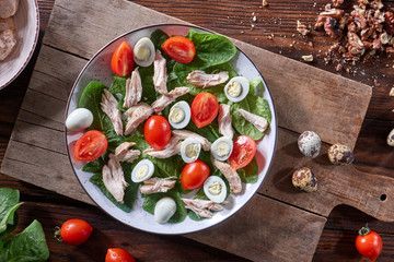 Healthy freshly prepared salad of quail eggs, meat, tomatoes and spinach in a plate on a wooden board on the kitchen table. Dietary lunch. Flat lay