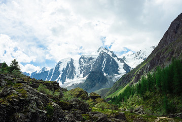 Snowy mountain top behind rocky mountains under cloudy sky. Rocky slope above forest. Clouds above glacier. Atmospheric landscape of majestic nature of highlands.