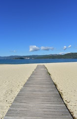 Beach in a bay with bright sand, blue water and wooden boardwalk. Galicia, Spain. Sunny day, blue sky.