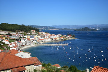 Coastal village in a bay with beach, boats and island with forest. Galicia, Rias Baixas, Spain. Sunny day, blue sky.