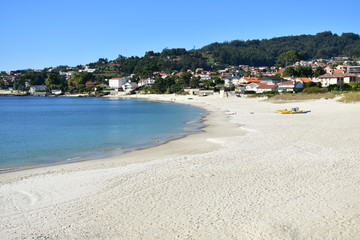 Beach with bright sand and turquoise water. Coastal village, trees and blue sky. Galicia, Rias Baixas, Spain.