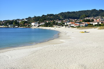 Fototapeta na wymiar Beach with bright sand and turquoise water. Coastal village, trees and blue sky. Galicia, Rias Baixas, Spain.