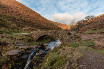 An autumnal waterfall and stone packhorse bridge at Three Shires Head in the Peak District.