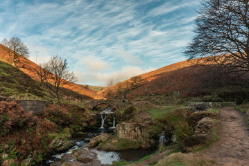 An autumnal waterfall and stone packhorse bridge at Three Shires Head in the Peak District.