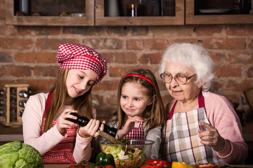 Two cute little sisters help her granny to prepare vegetable salad at kitchen at home