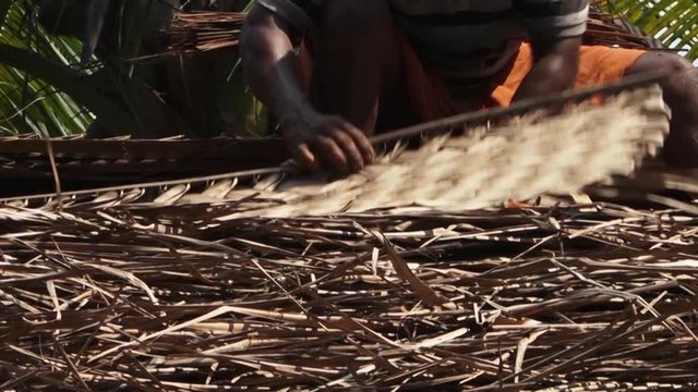 A low-angle, close-up shot of a man placing braided leaves on a thatch roof.
