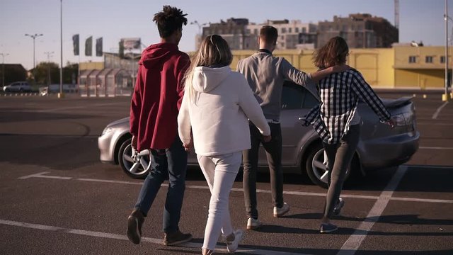 Rare view of a multirace trendy young two couples walking by mall parking zone and sit into the silver coloured car. Two couple having time together