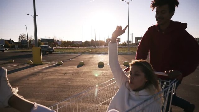 Happy young couple riding on trolley on empty mall parking , hipster friend have good time after shopping, couple in love riding on shopping cart, multirace. Sun shines on the background