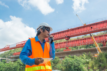 Architect or Engineer, wearing white Hardhat and safety vest, use Radio Communication to supervise...