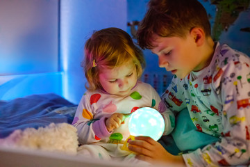Cute little toddler girl and kid boy playing with colorful night light lamp before going to bed. Sleepy tired baby sister and brother in nightwear having fun. Healthy children, siblings not sleeping.