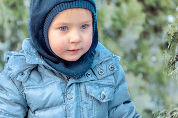 Cute caucasian liittle boy with bright blue eyes in winter clothes and hat (hood) on blue pine and fir background. Healthy childhood. Outdoors, copy space, close up portrait.