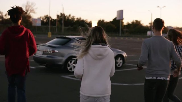 Rare view of a multirace trendy young two couples running by mall parking zone and sit into the silver coloured car. Two couple having time together. Evening dusk