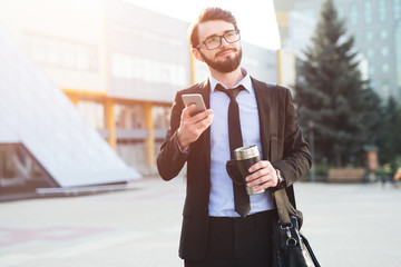 Hipster impressed businessman using smartphone on office build background with coffee thermo mug in hand and black bag.