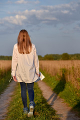 Girl walking on road among wheat field at dawn in jeans and a white shirt photos scatter in hand profoundly sad, light, letting go past