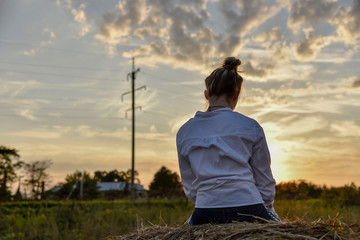 Girl with beautiful hair at sunset sitting on a haystack back to the photographer