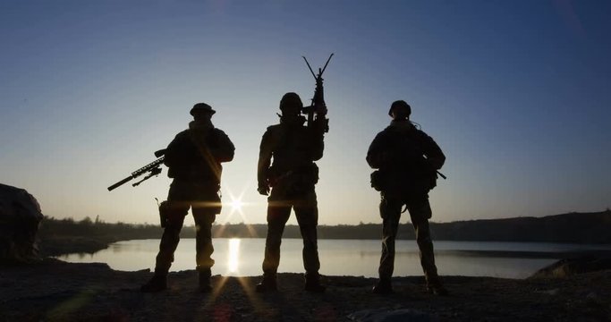 Medium shot of three soldiers posing with guns against the backdrop of a lake as the sun sets behind them