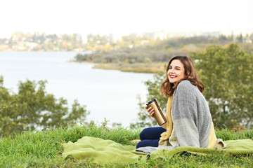 Naklejka na ściany i meble Beautiful young woman with thermos resting on plaid outdoors