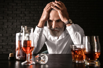 Depressed senior man drinking whiskey at table on dark background