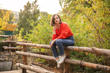 Naklejka na ściany i meble Beautiful fashionable woman sitting on wooden fence outdoors