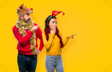 Couple dressed up for the christmas holidays pointing finger to the side with a surprised face on yellow background