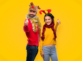 Couple dressed up for the christmas holidays giving a thumbs up gesture and smiling on yellow background
