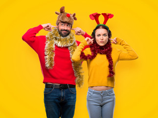 Couple dressed up for the christmas holidays covering ears with hands on yellow background