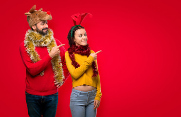 Couple dressed up for the christmas holidays pointing to the side with a finger to present a product on isolated red background