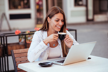 Smiling young woman looking at laptop while drinking coffee in outdoor cafe.